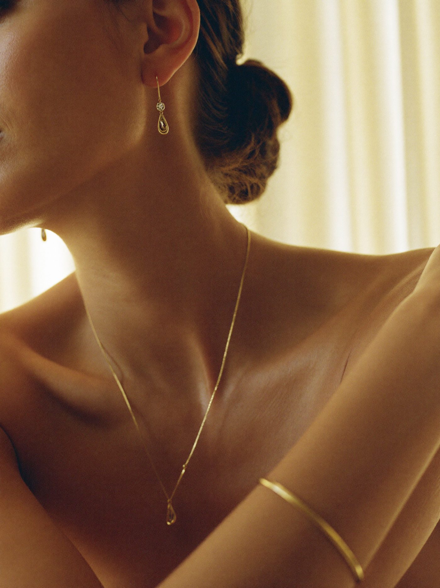 Detalhe de brincos de ouro com cristais, colar fino e pulseira dourada em uma mulher de ombros à mostra