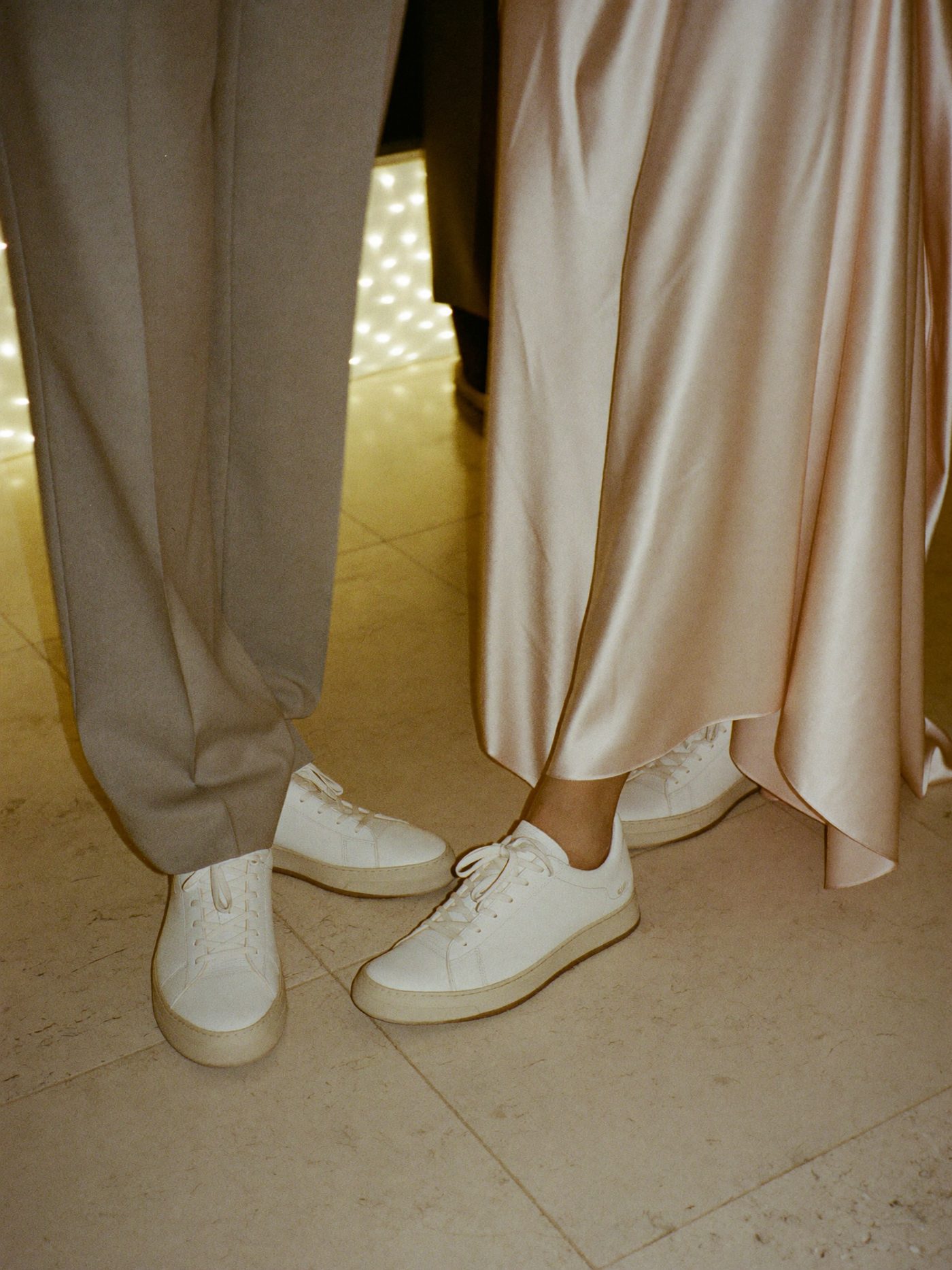 Casal de convidados com tênis brancos: ele de calça social cinza, ela de vestido longo rosé