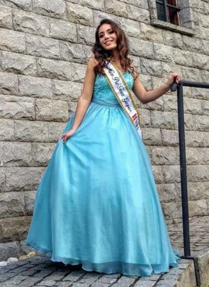 Luane Silveira de vestido longo azul-claro e faixa de Miss Pré-Teen Gaspar, na escadaria de pedra