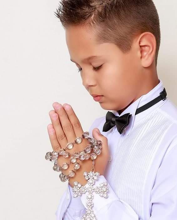 Menino de camisa branca e gravata-borboleta segurando um terço com as mãos em oração